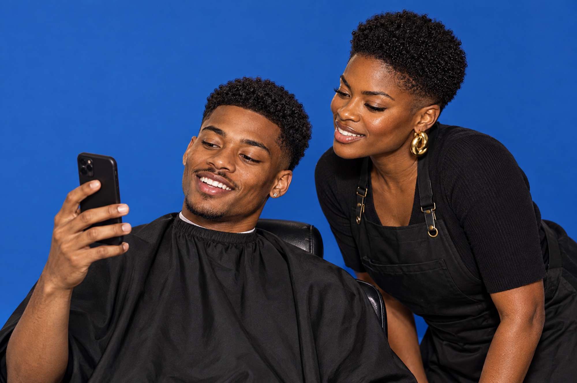 A client in a barber chair shows his barber a reference photo on his phone; she leans in, both smiling, against cobalt paper.