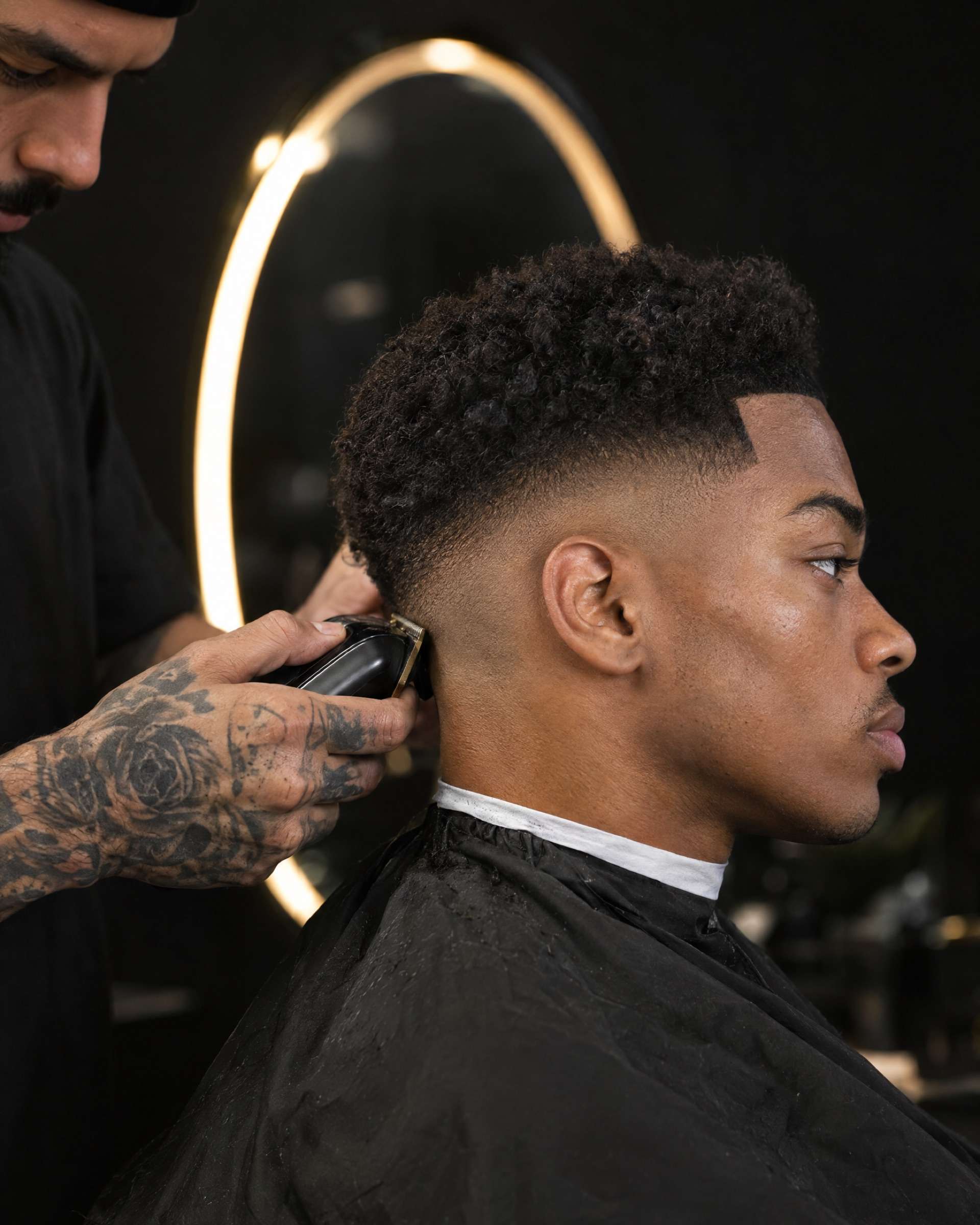 Primer plano: la mano tatuada de un barbero termina un skin fade con máquina en un joven de perfil, con un espejo ovalado iluminado detrás.
