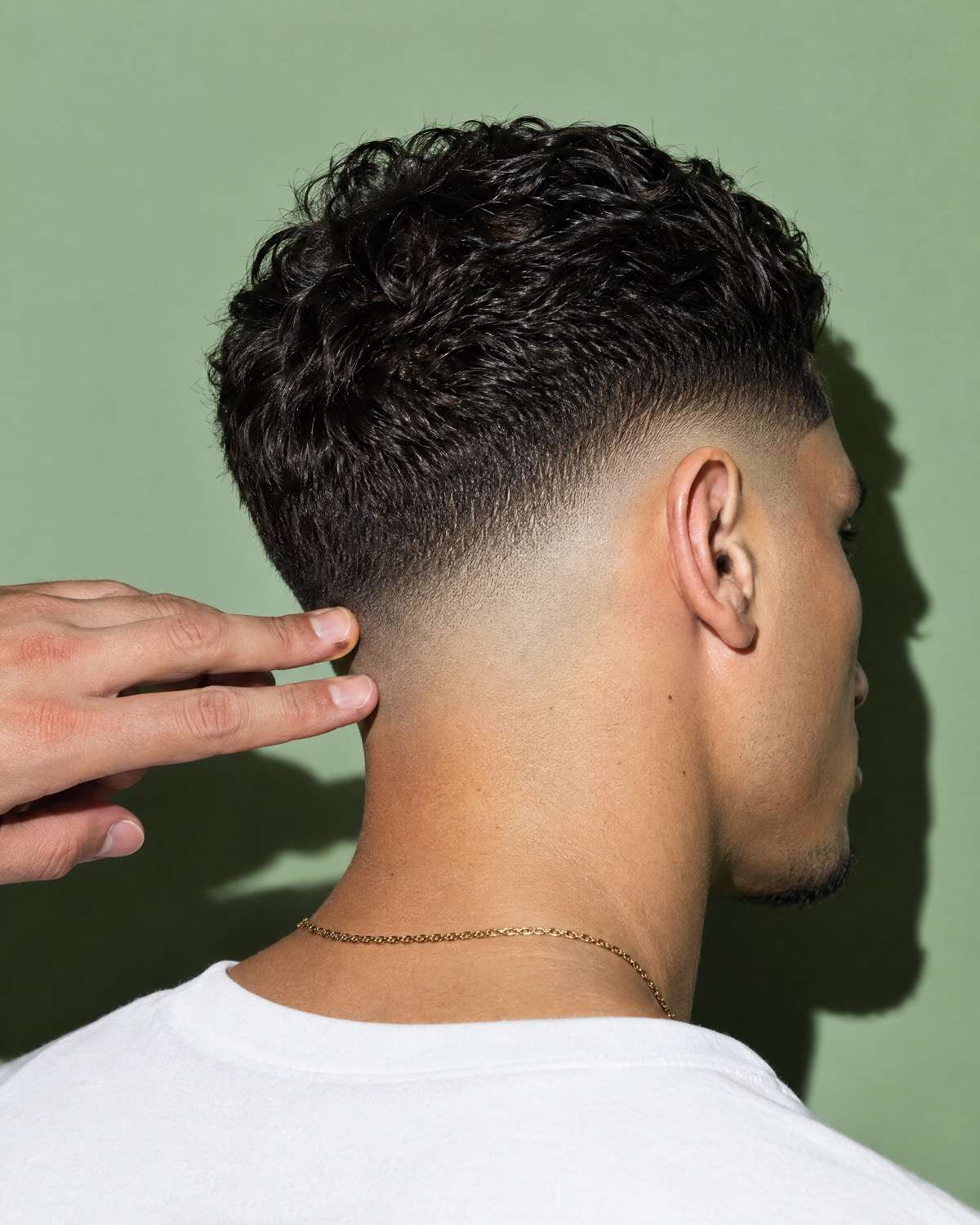 La nuca de un fade perfecto sobre papel salvia, con los dedos del barbero revisando la línea.