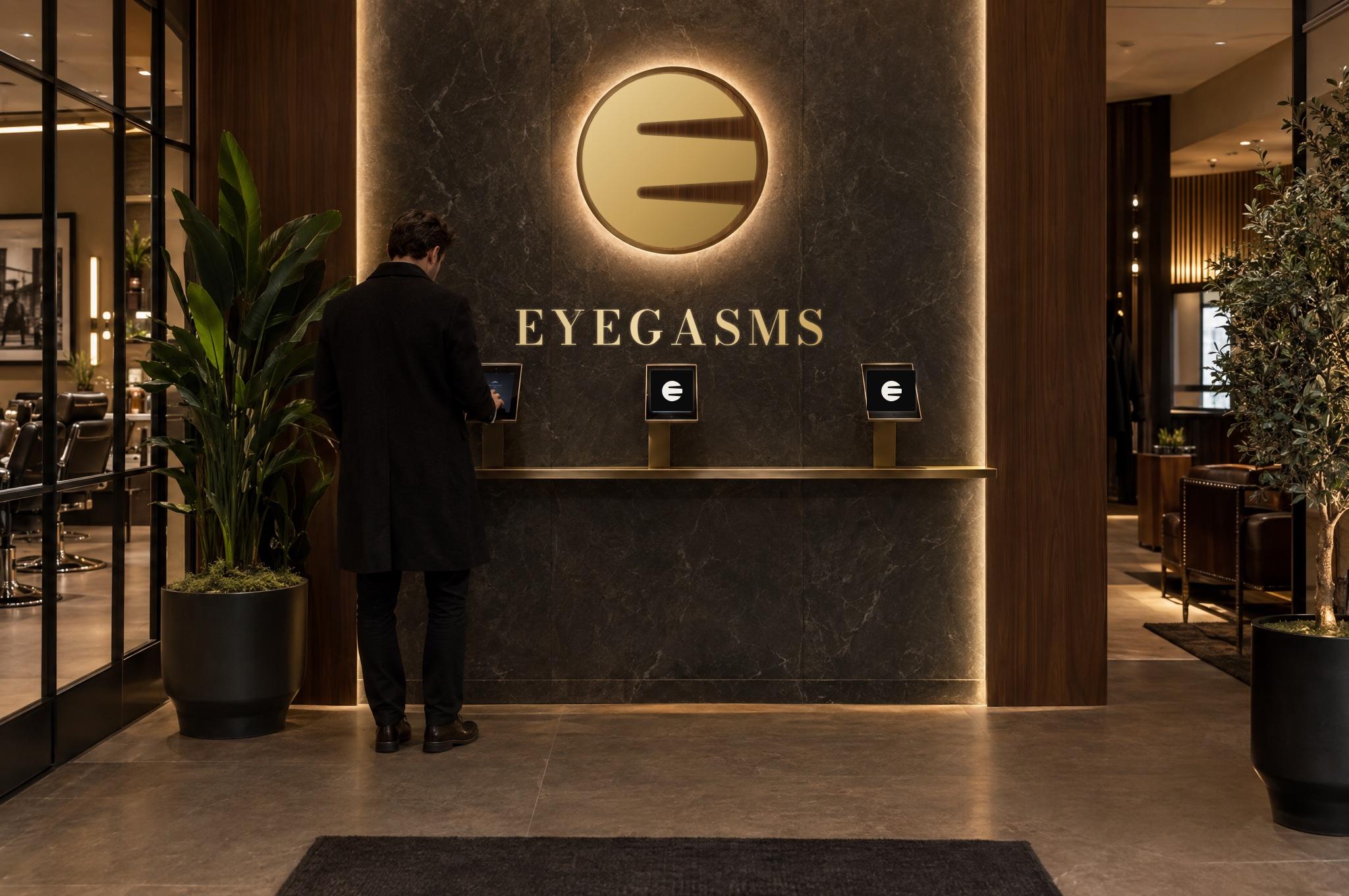 El muro de check-in frente a la entrada: piedra oscura con la E de EYEGASMS en latón brillando sobre un disco iluminado, el nombre EYEGASMS en latón debajo y tres estaciones iPad con la E sobre una repisa de latón; un hombre hace check-in.
