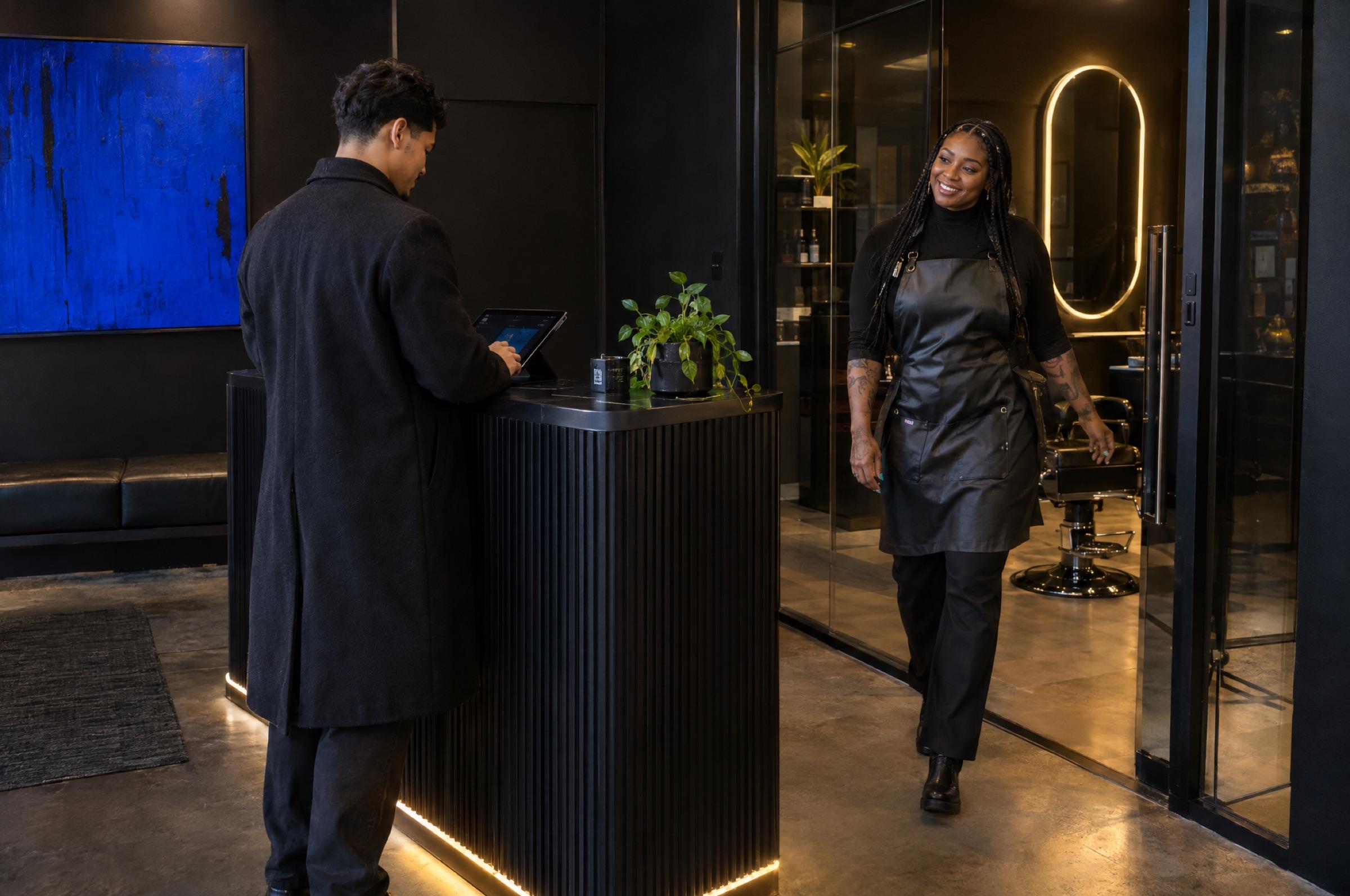 Arrival: a man checks in on a tablet at a black ribbed desk with a line of light at its base while a smiling barber with braids and tattoos steps out of her glass studio to greet him; a cobalt painting hangs behind.