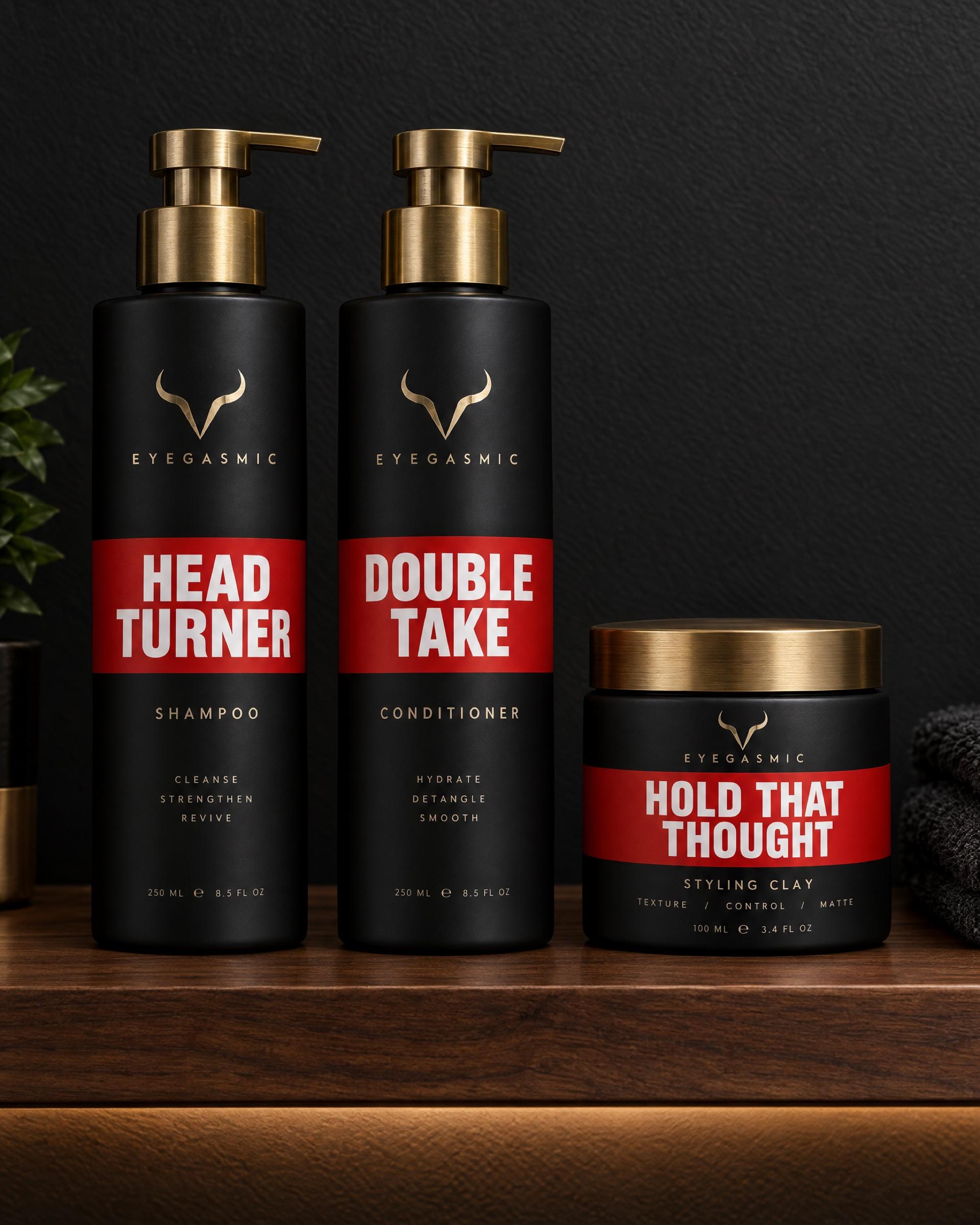EYEGASMIC hair care in signal red on a lit walnut shelf: Head Turner shampoo, Double Take conditioner and Hold That Thought styling clay.