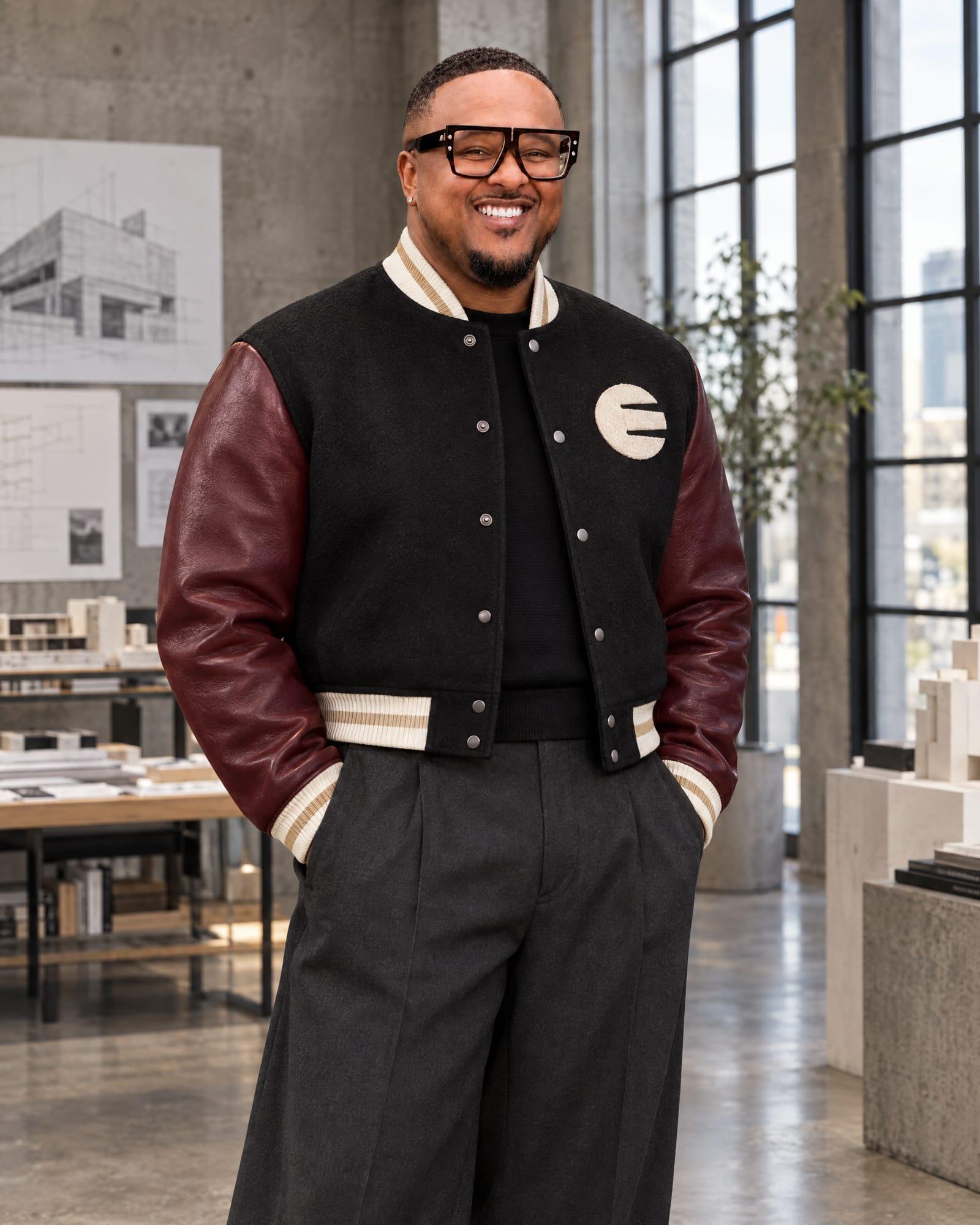 Brennon Jackson, founder of EYEGASMS, smiling in glasses in a concrete architecture studio, wearing the founder's edition of the EYEGASMS architectural letterman: black wool body, oxblood leather sleeves, bone trims with a champagne stripe and a bone chenille circle-E patch, over a black knit with wide-leg charcoal trousers.