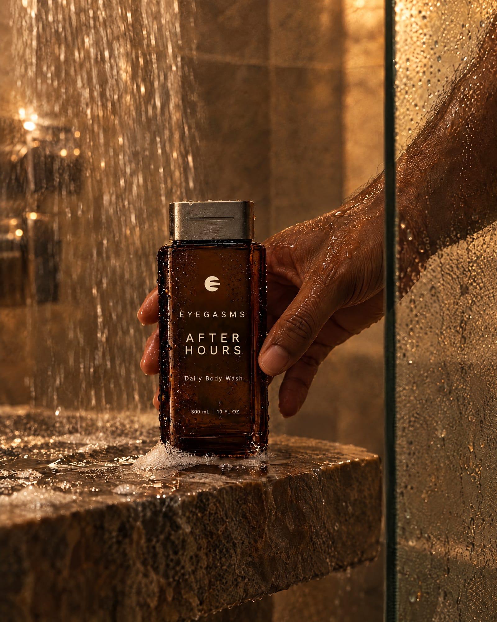 After Hours, daily body wash, in EYEGASMS E-Form packaging: a man's hand lifts the smoked espresso bottle from a wet stone shower ledge.