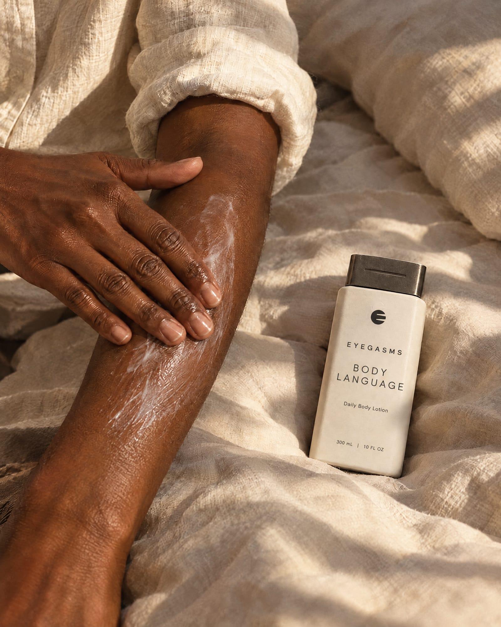Body Language, daily body lotion, in EYEGASMS E-Form packaging: hands smooth lotion along a forearm on linen; the bone bottle lies beside them.