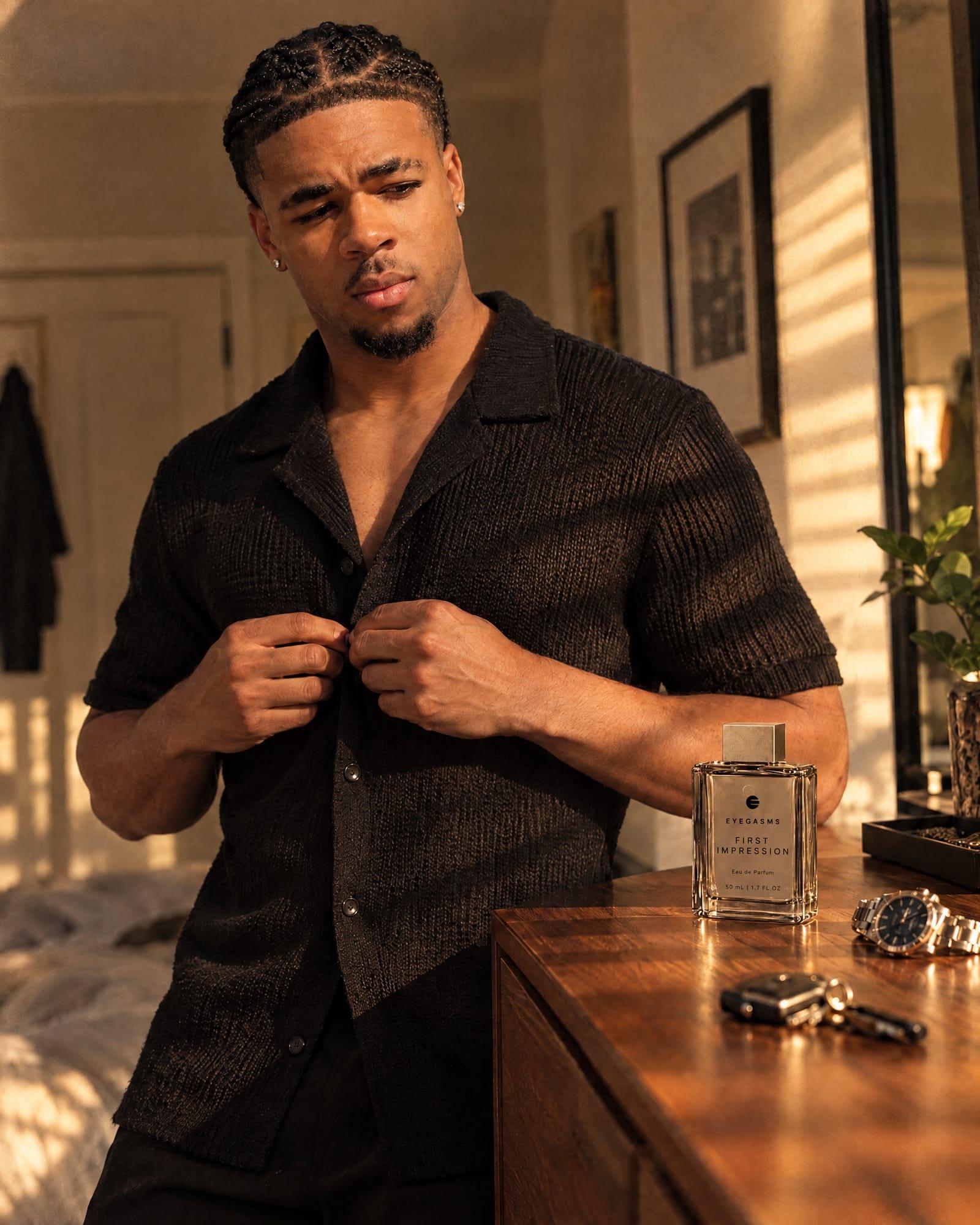 First Impression, eau de parfum, in EYEGASMS E-Form packaging: a young Black man with braids buttons a black knit shirt beside the clear flacon on a walnut dresser.