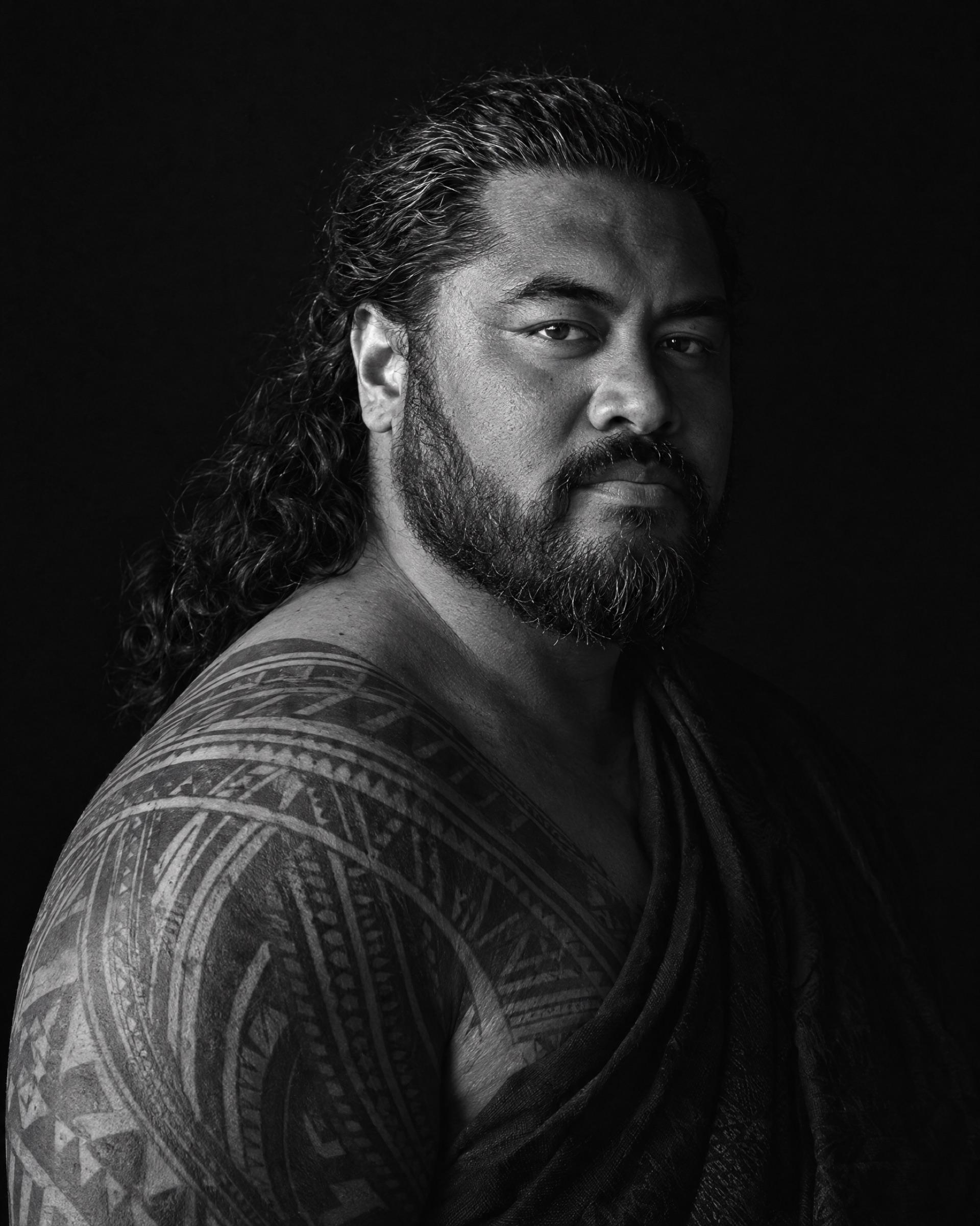 Black-and-white portrait of a man with long hair pulled back, a full beard and a shoulder tattoo.