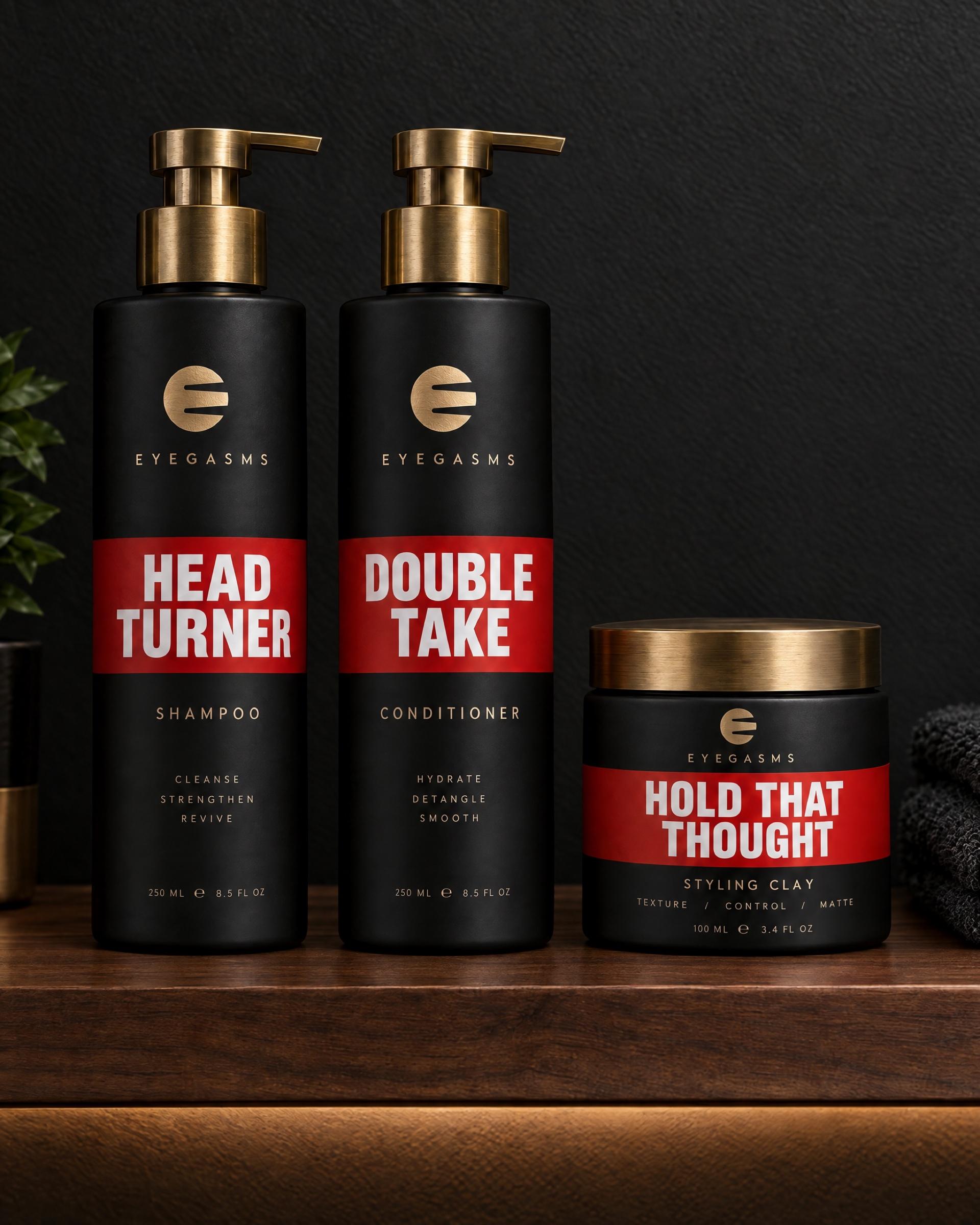 Cuidado del cabello EYEGASMS en rojo sobre una repisa de nogal iluminada: shampoo Head Turner, acondicionador Double Take y arcilla Hold That Thought.